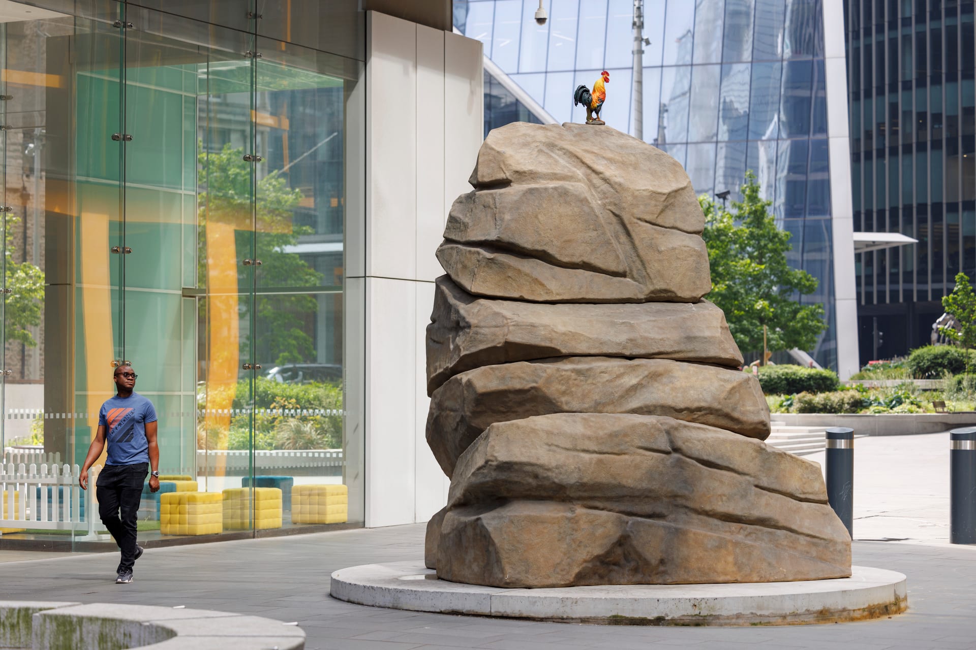 A large sculpture of a boulder with a cockerel on the top situated in a square surrounded by glass buildings 