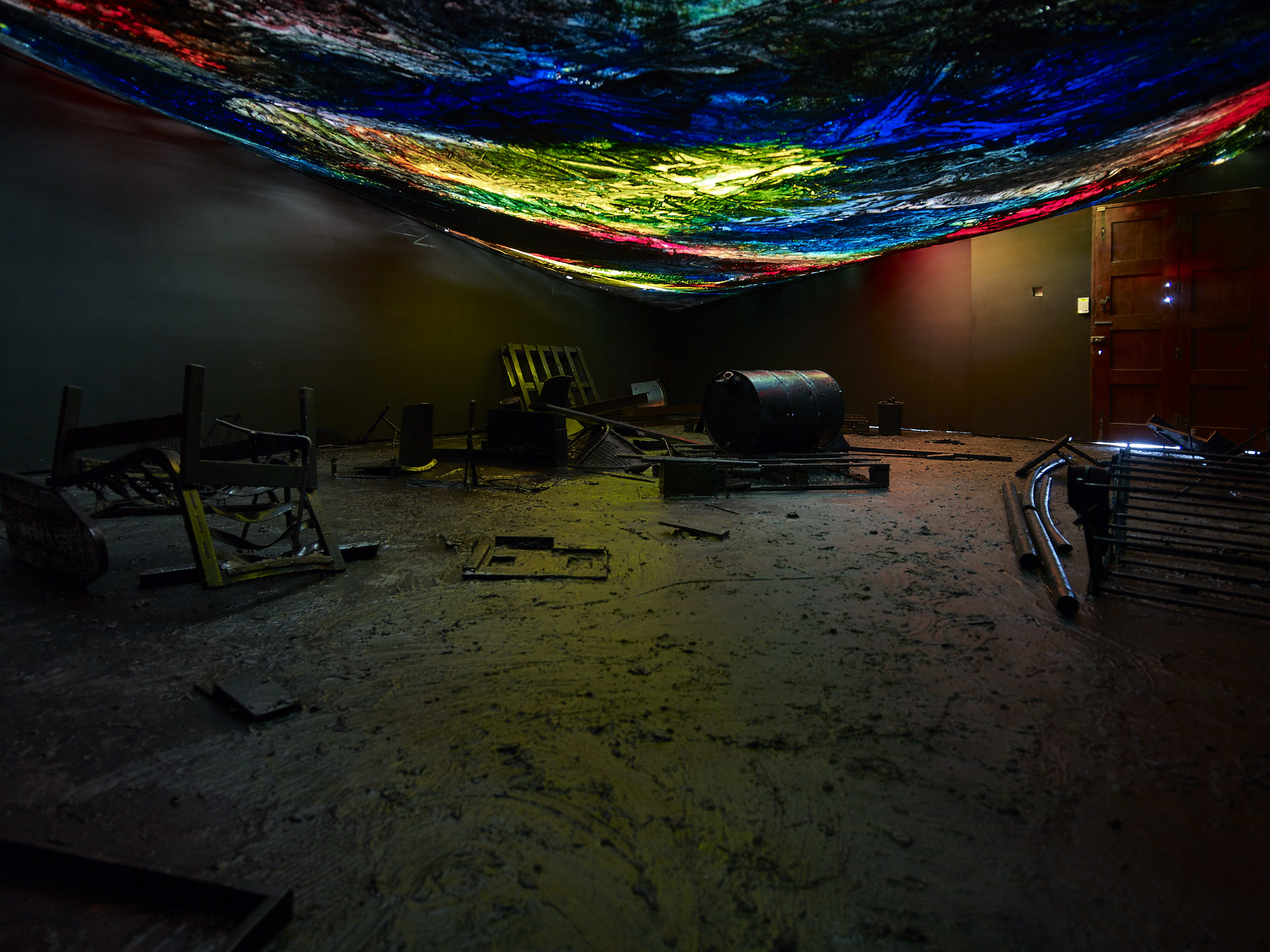 A installation by Hugo Canoilas consisting of darkened room with objects covered in black latex, and a low painted colourful canvas as ceiling