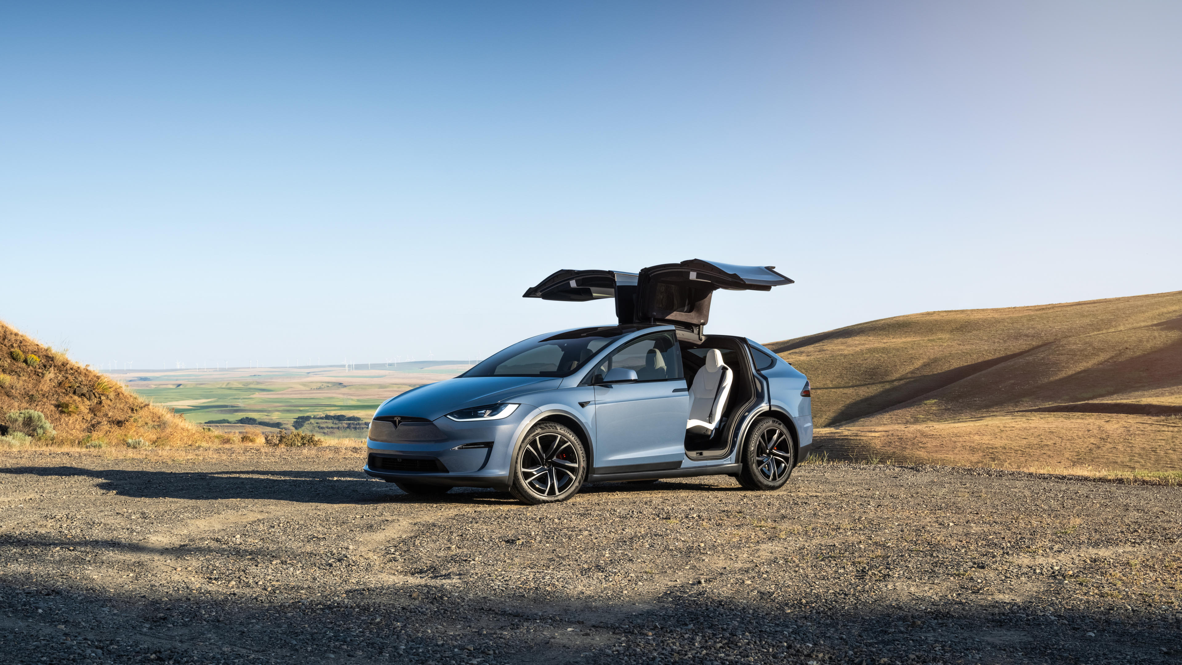 A light blue Tesla Model X with its falcon wing doors open, parked on a gravel path overlooking rolling hills.