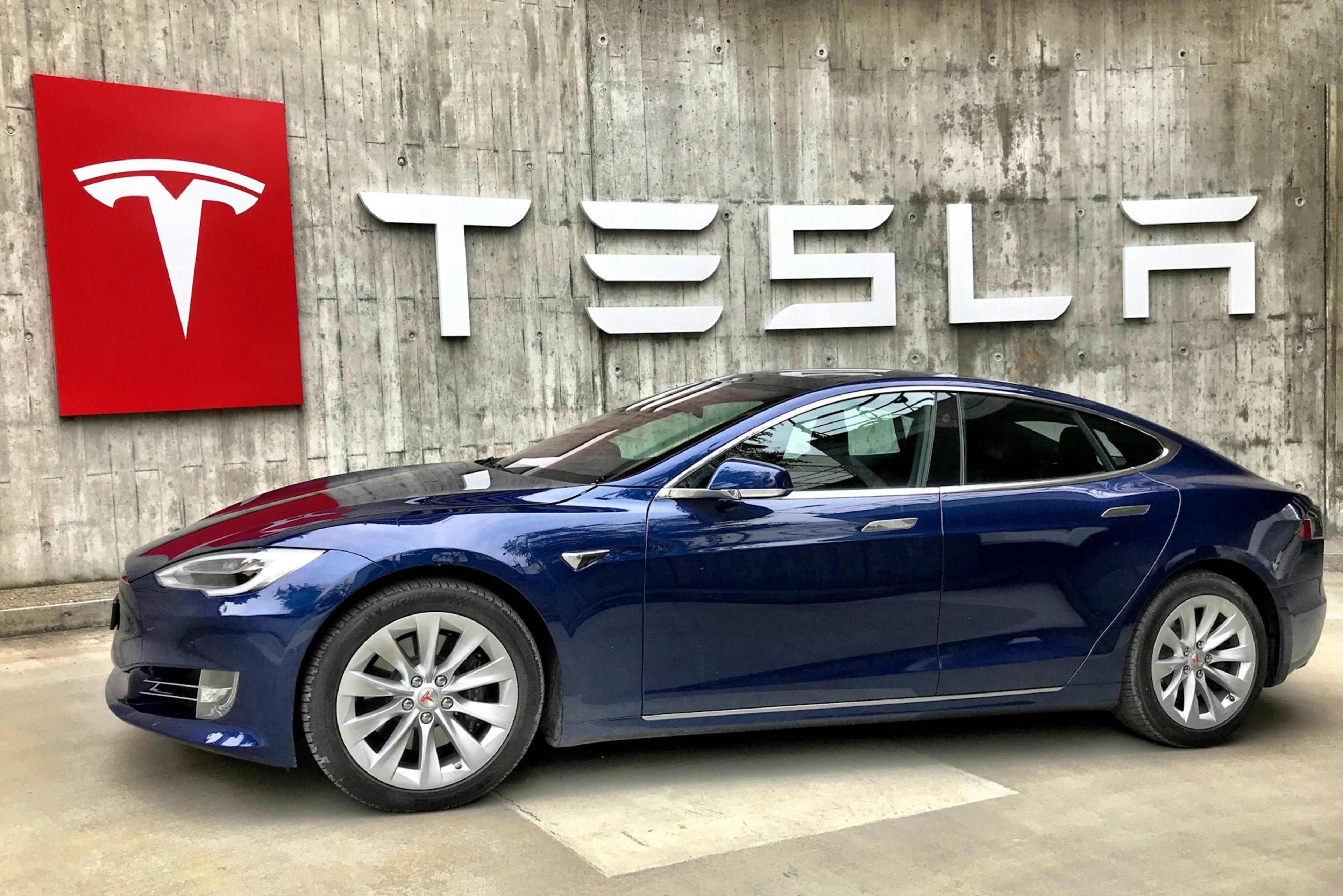 A blue Tesla car parked in front of a Tesla logo sign on a wall