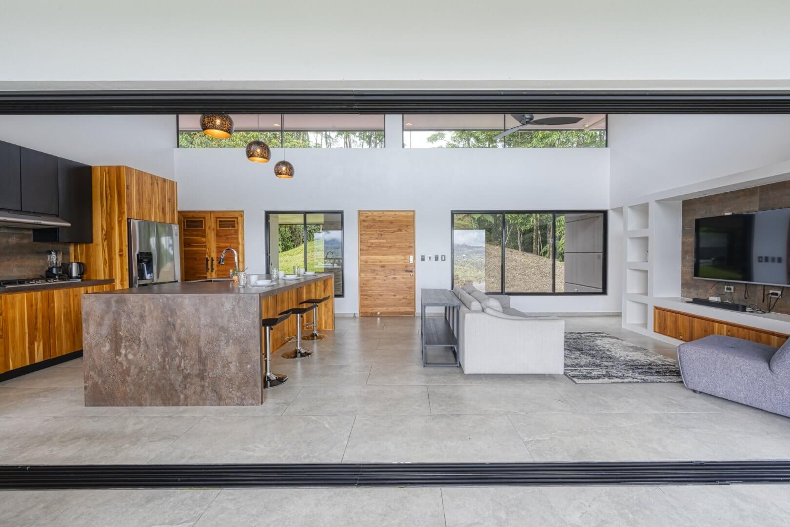 Interior view of a luxury modern home's open-concept kitchen and living room, featuring a large island, wooden cabinetry, comfortable seating, and wide sliding doors opening to jungle views in Escaleras, Dominical.