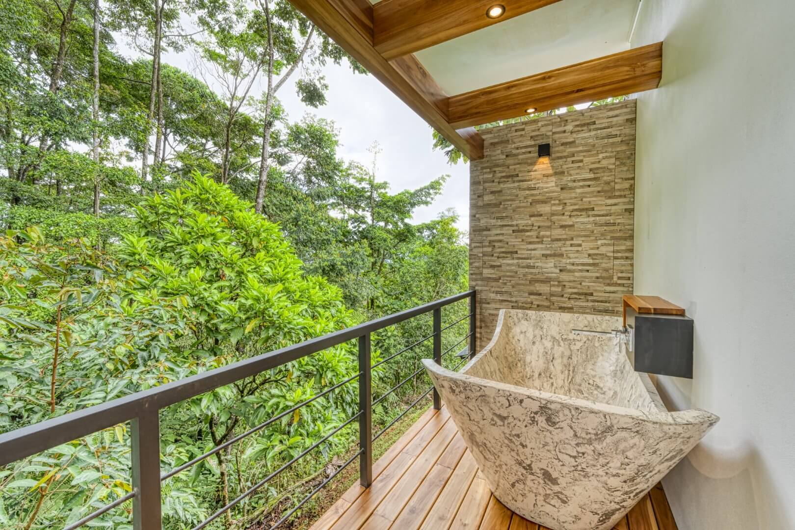 An outdoor bathtub made of light-colored stone, situated on a wooden deck with a metal railing, overlooking a dense green jungle canopy under a cloudy sky.