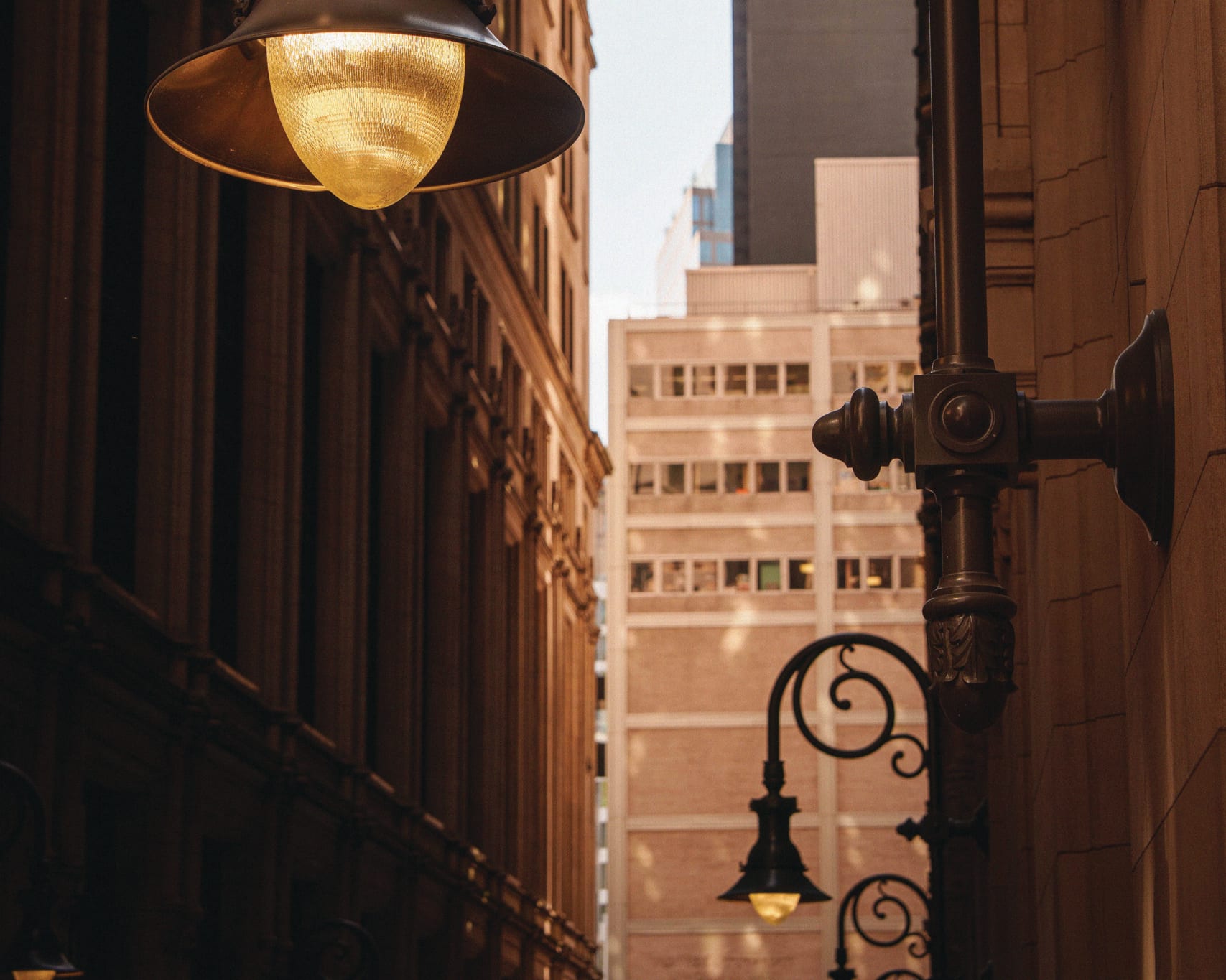 Two ornate, dark bronze streetlights with glowing yellow lamps. One is large and prominent in the foreground, while a smaller one is visible in the background. Tall city buildings and a sliver of sky are visible between them.