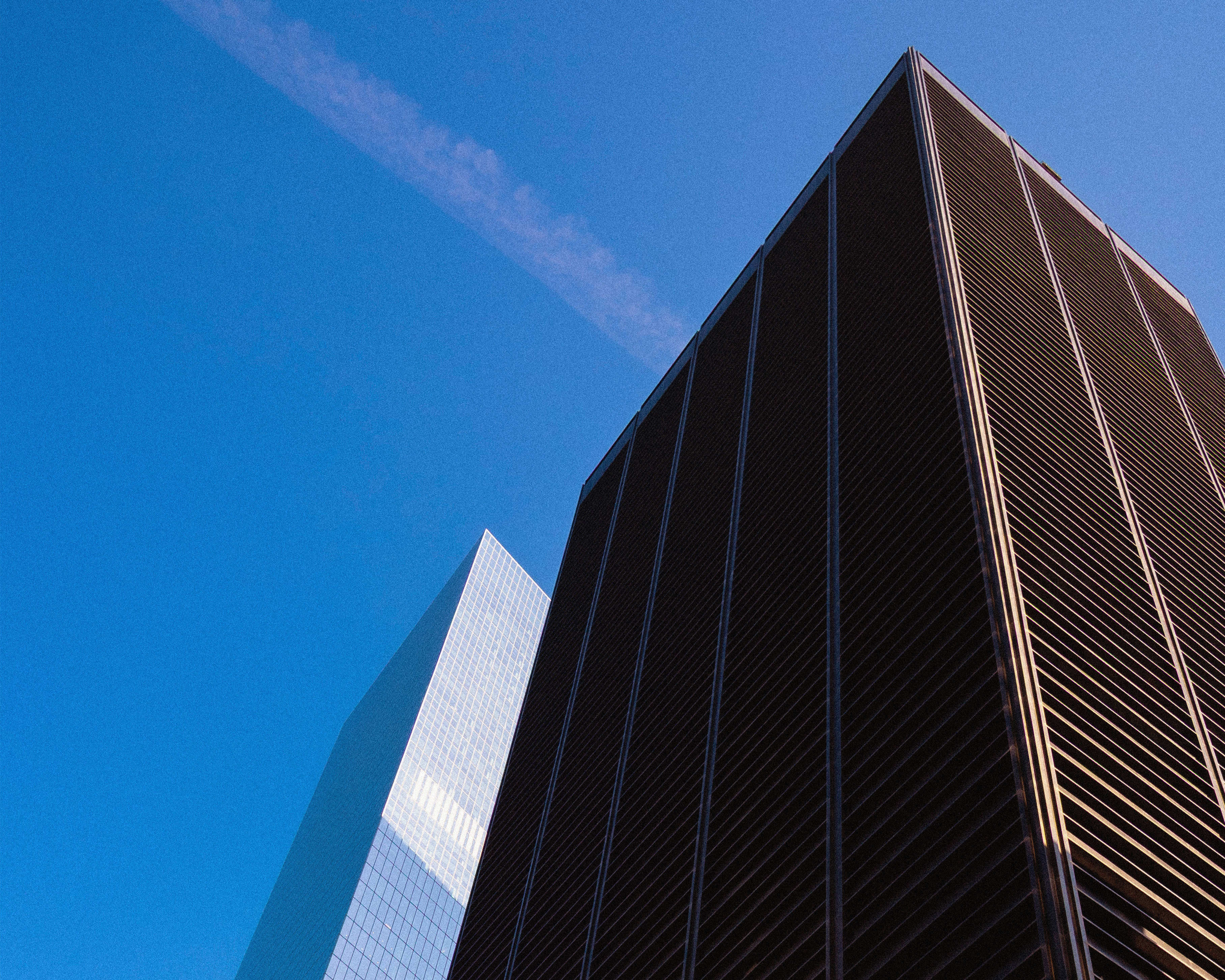 Two tall buildings against a blue sky. The building on the right is dark with horizontal lines, and the building on the left is reflective glass.