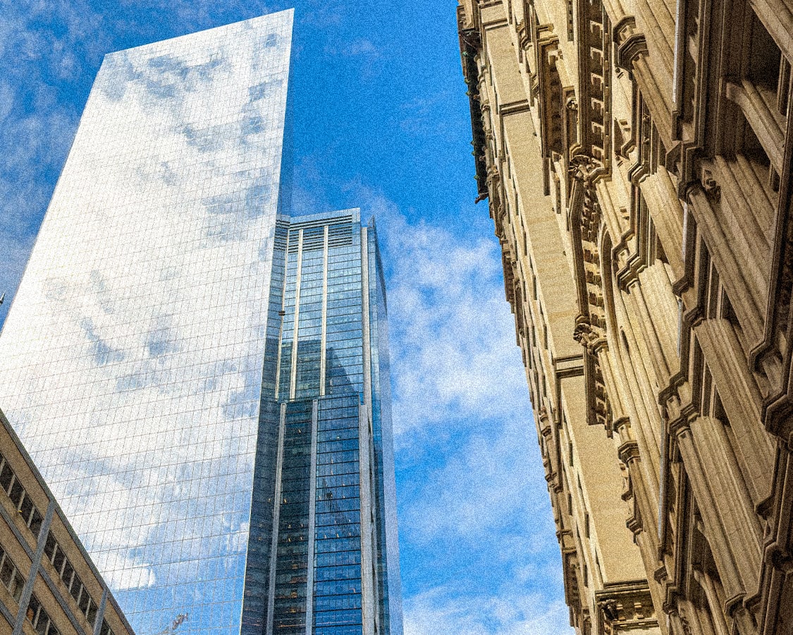 Low-angle view of multiple skyscrapers against a blue sky. A large, reflective glass building is central, flanked by older, ornate buildings and another modern glass tower.