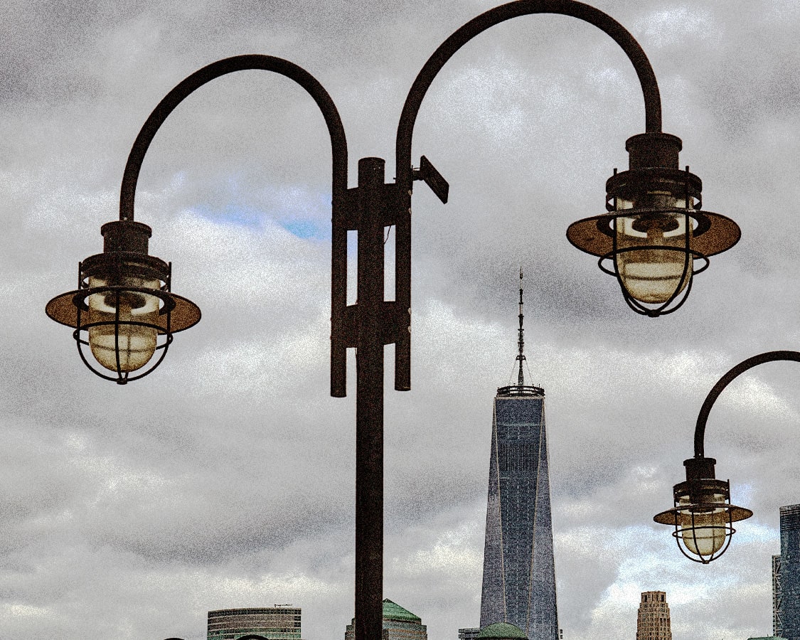 Several dark, arched streetlights with caged lanterns in the foreground, framing the One World Trade Center skyscraper and other city buildings under a cloudy sky.