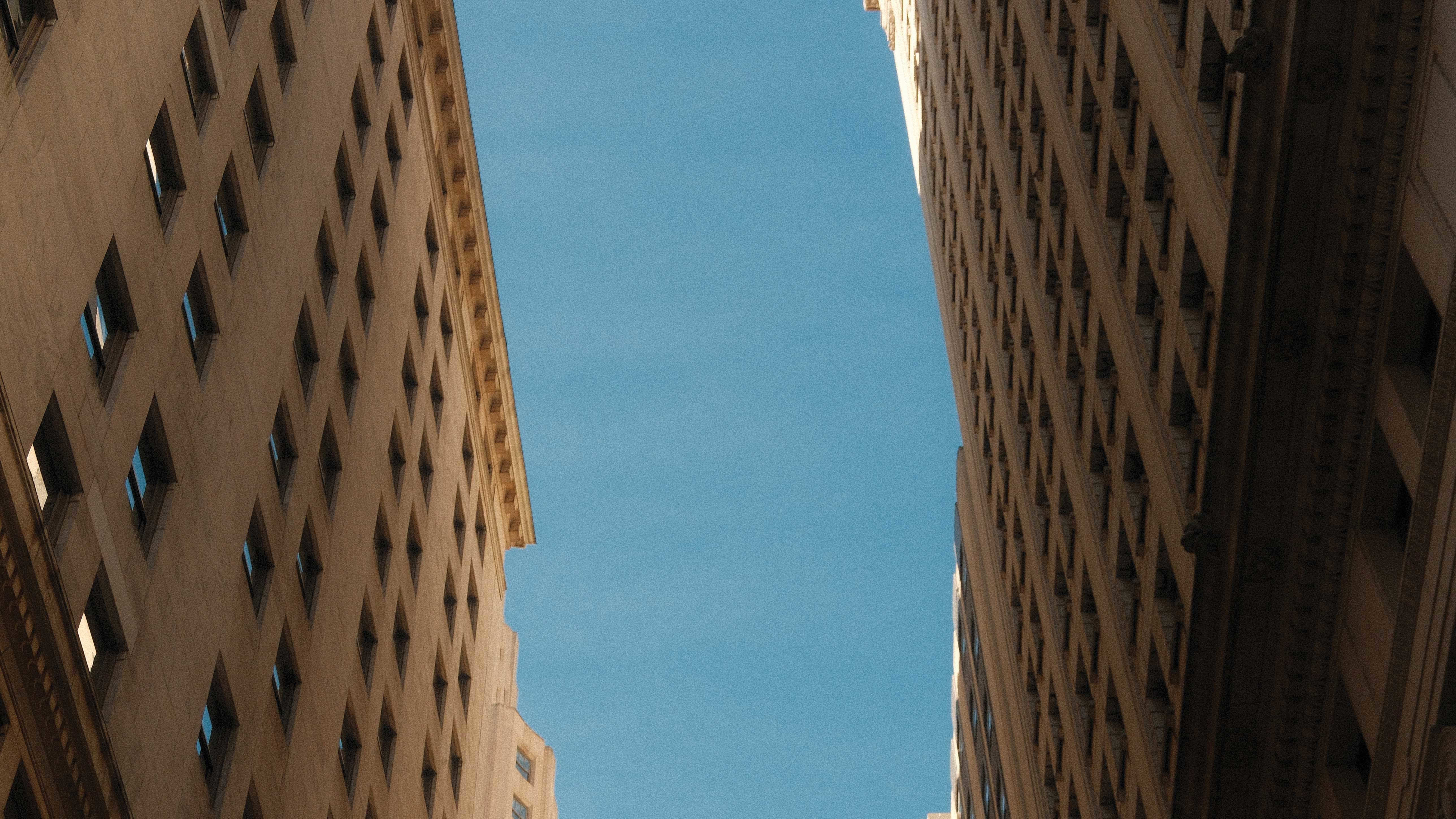 Low-angle view of tall, light-colored buildings framing a clear blue sky, with a dark church spire and modern glass buildings visible in the background.