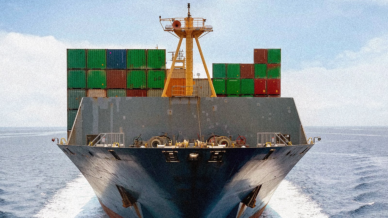 Front view of a large container ship on the ocean, laden with green and red shipping containers, leaving a white wake.