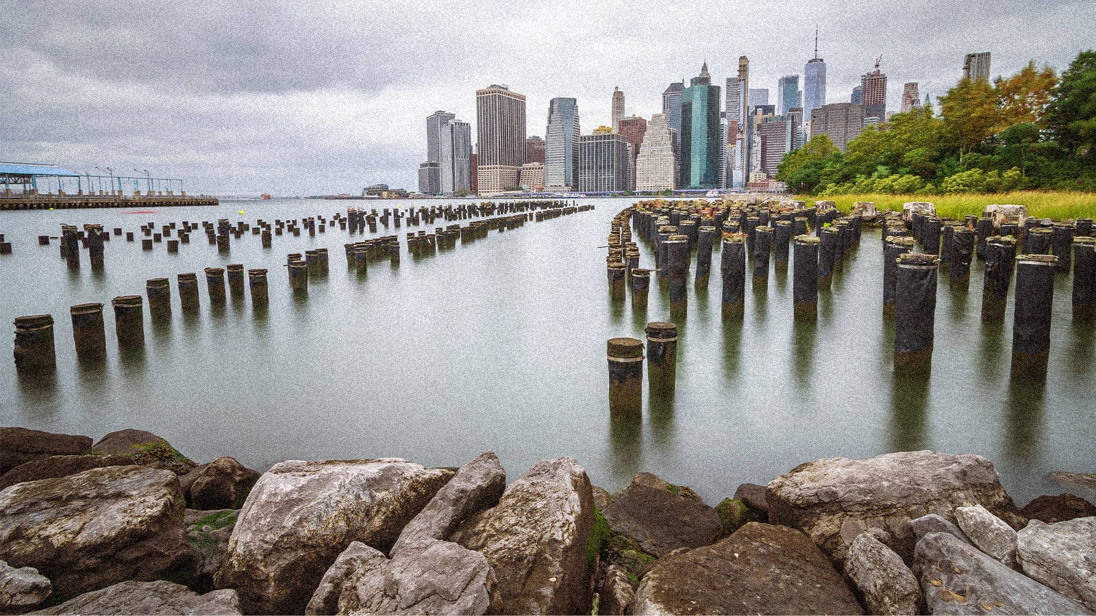 A wide shot of a city skyline with numerous dark wooden pilings in the water and a rocky shore in the foreground, under a gray, cloudy sky.