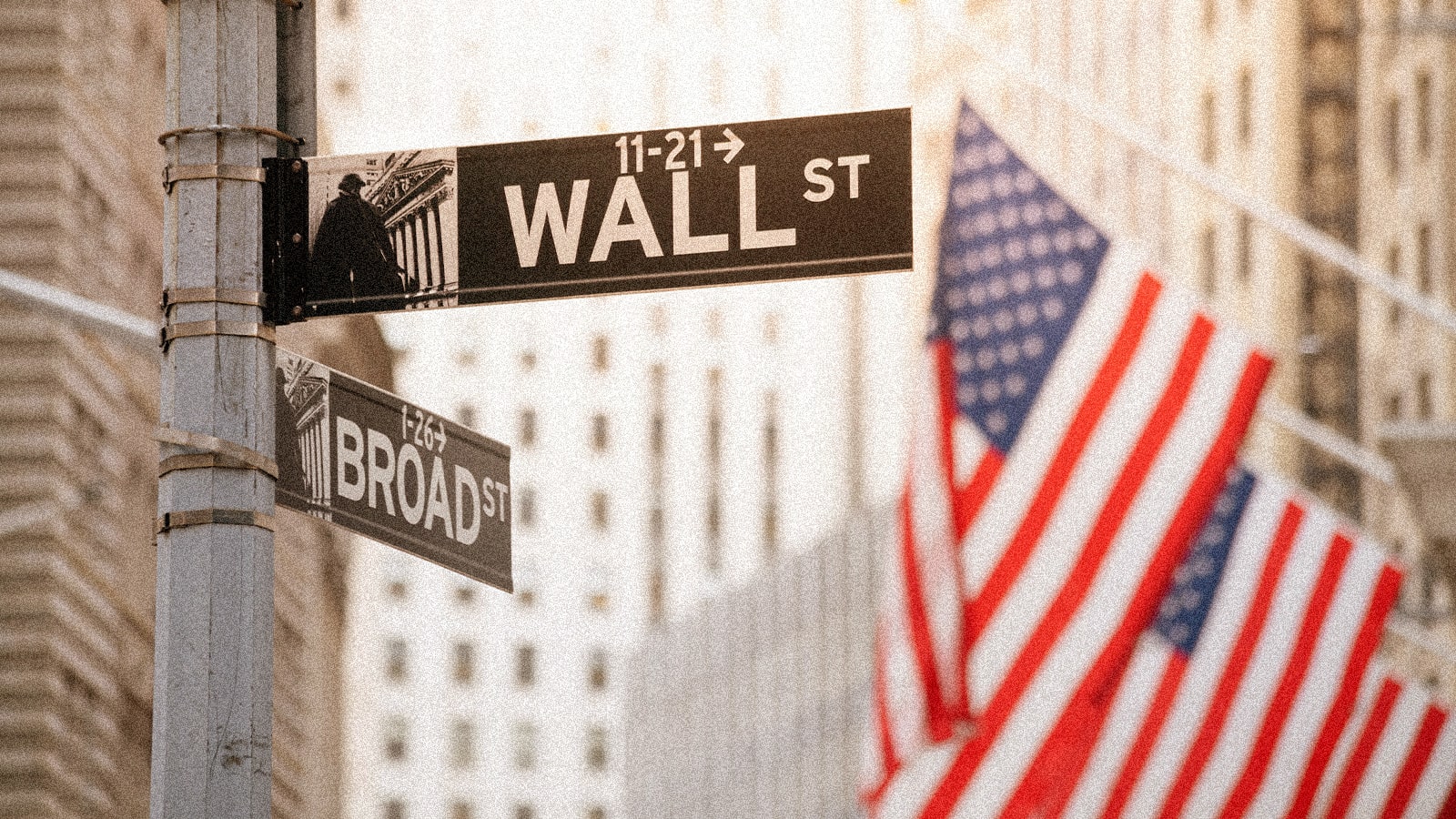 Street signs for Wall Street and Broad Street on a metal pole, with American flags and blurred buildings in the background.