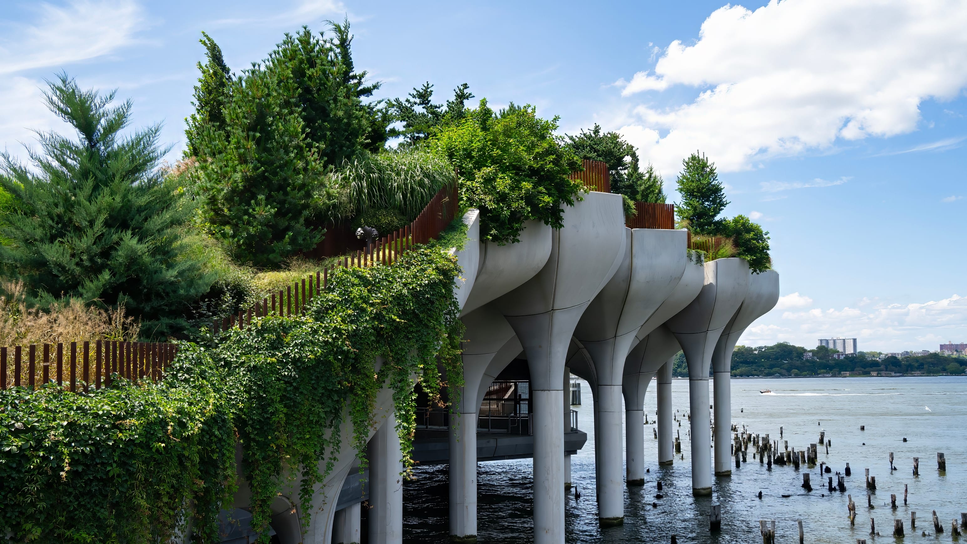 An elevated park on tulip-shaped concrete supports rises from the Hudson River, covered in green trees and vines, with old wooden pilings in the water and a distant city skyline under a blue sky.
