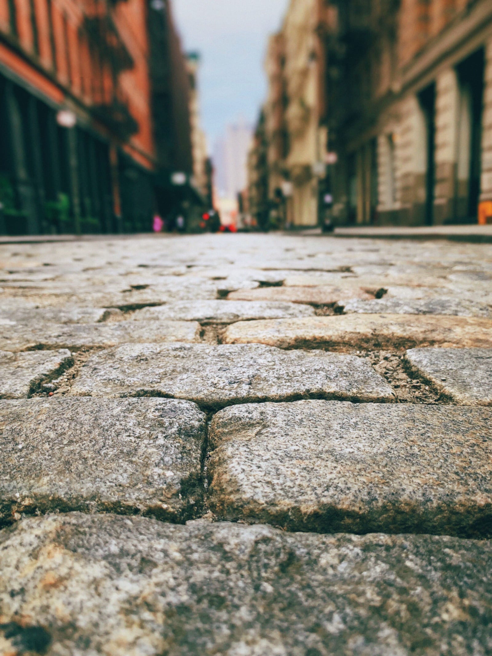 A low-angle view of a cobblestone street, sharply focused in the foreground, with blurred historic buildings lining both sides of the street in the background.