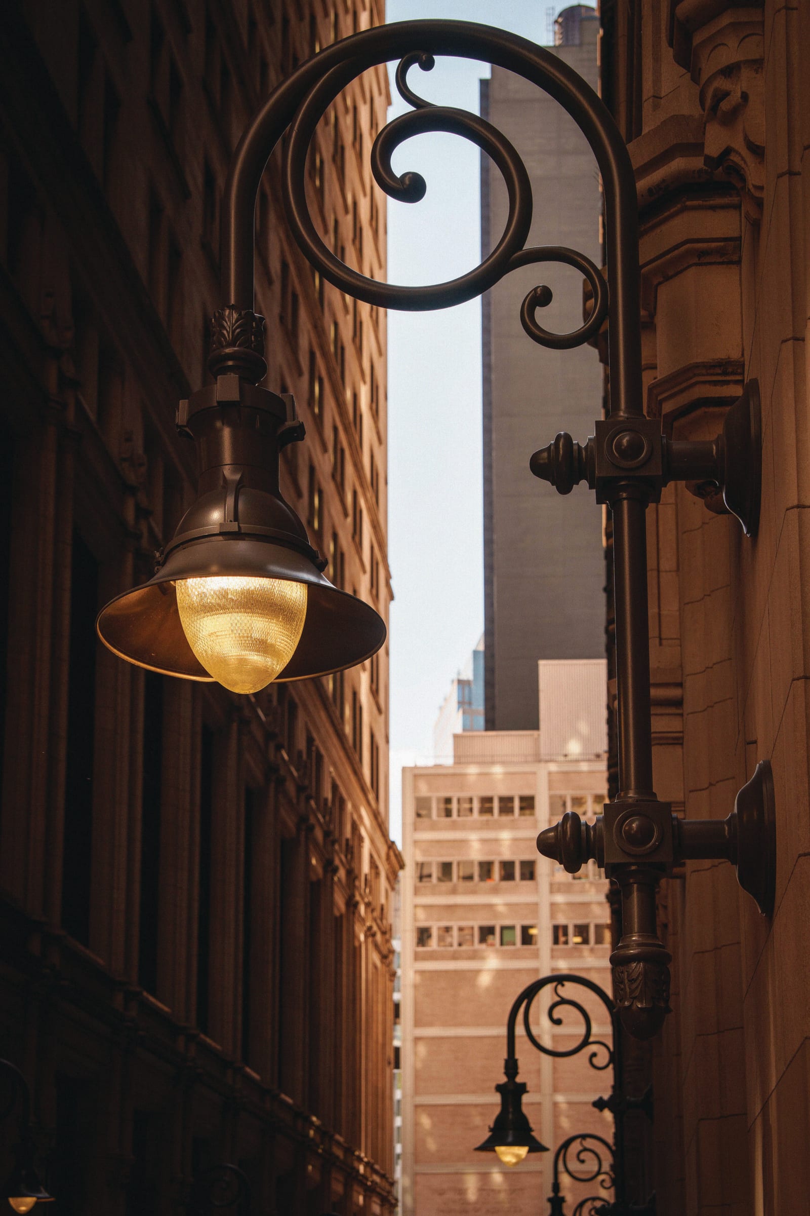 Two ornate, dark bronze streetlights with glowing yellow lamps. One is large and prominent in the foreground, while a smaller one is visible in the background. Tall city buildings and a sliver of sky are visible between them.