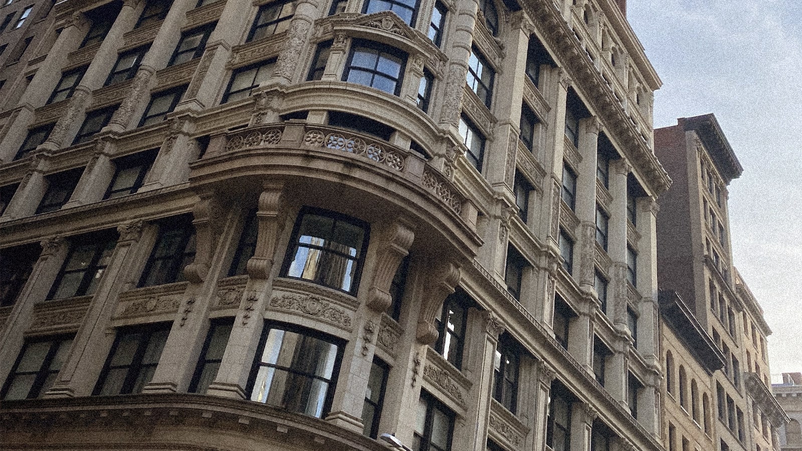 Low-angle view of an ornate, light-colored building with a curved corner, decorative balcony, and many windows, set against an overcast sky.