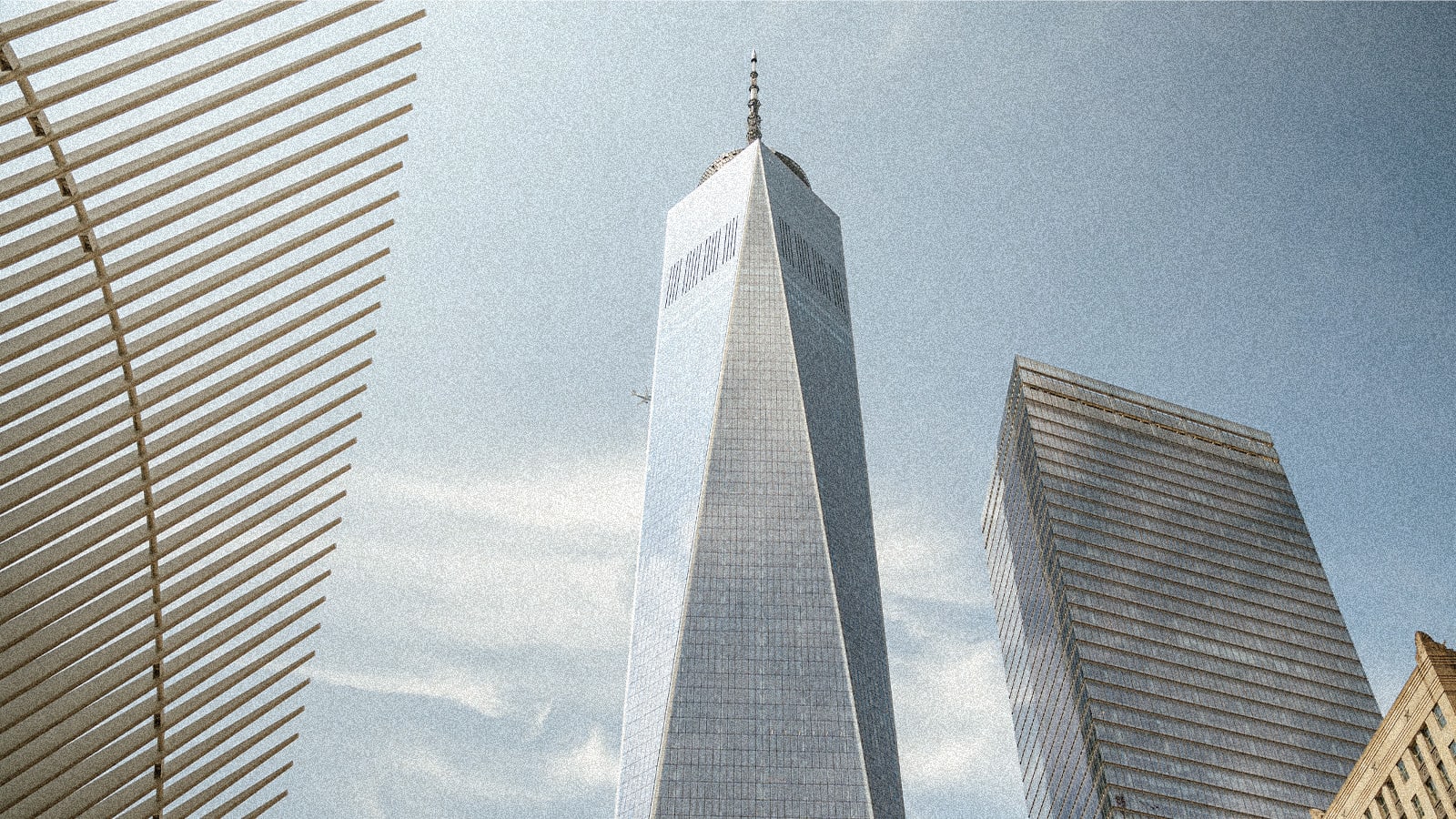 Low-angle view of One World Trade Center, the Oculus structure, and another glass skyscraper against a hazy blue sky.