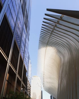 Low-angle view of a blue glass skyscraper on the left and the white, ribbed structure of the Oculus on the right, against a clear blue sky.