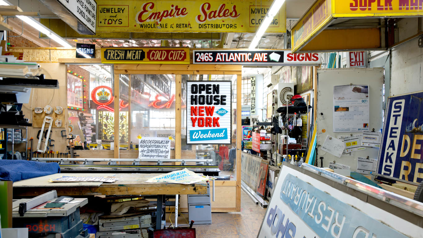 A cluttered sign shop interior features tools, worktables, and colorful vintage signs, including one reading Open House New York Weekend on a glass door surrounded by various business signs and advertisements.