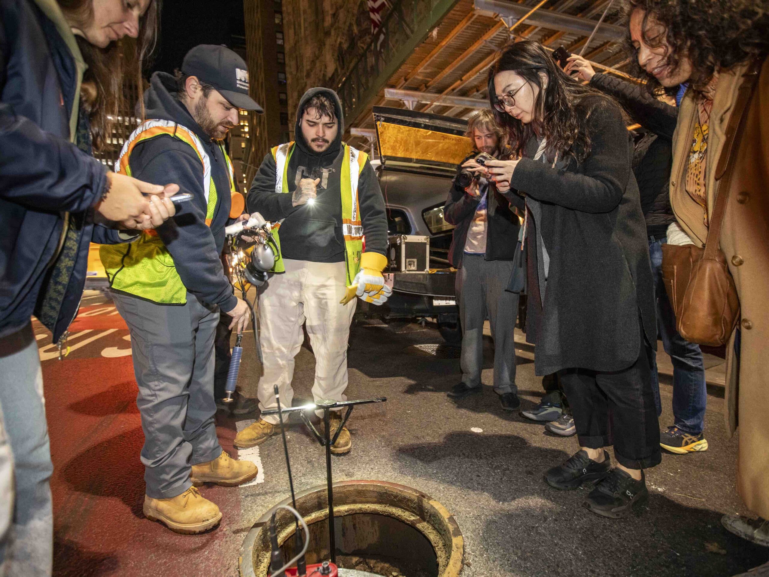 Two DEP workers demonstrate listening equipment beside an open manhole. A group of visitors watch them work.