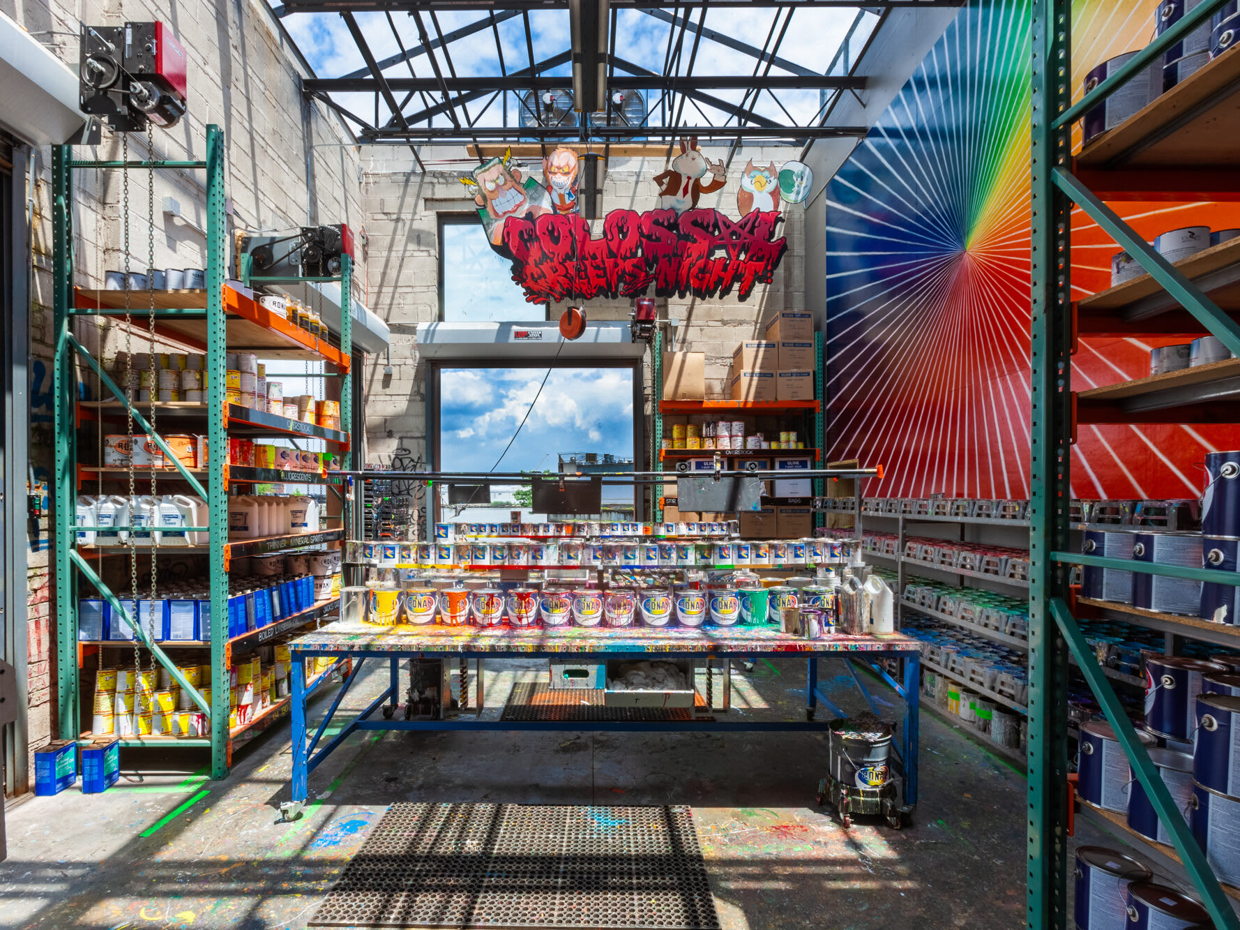 A paint shop with cans of paint lining the walls surrounding a central work table. Colorful murals are on the walls above the shelves.