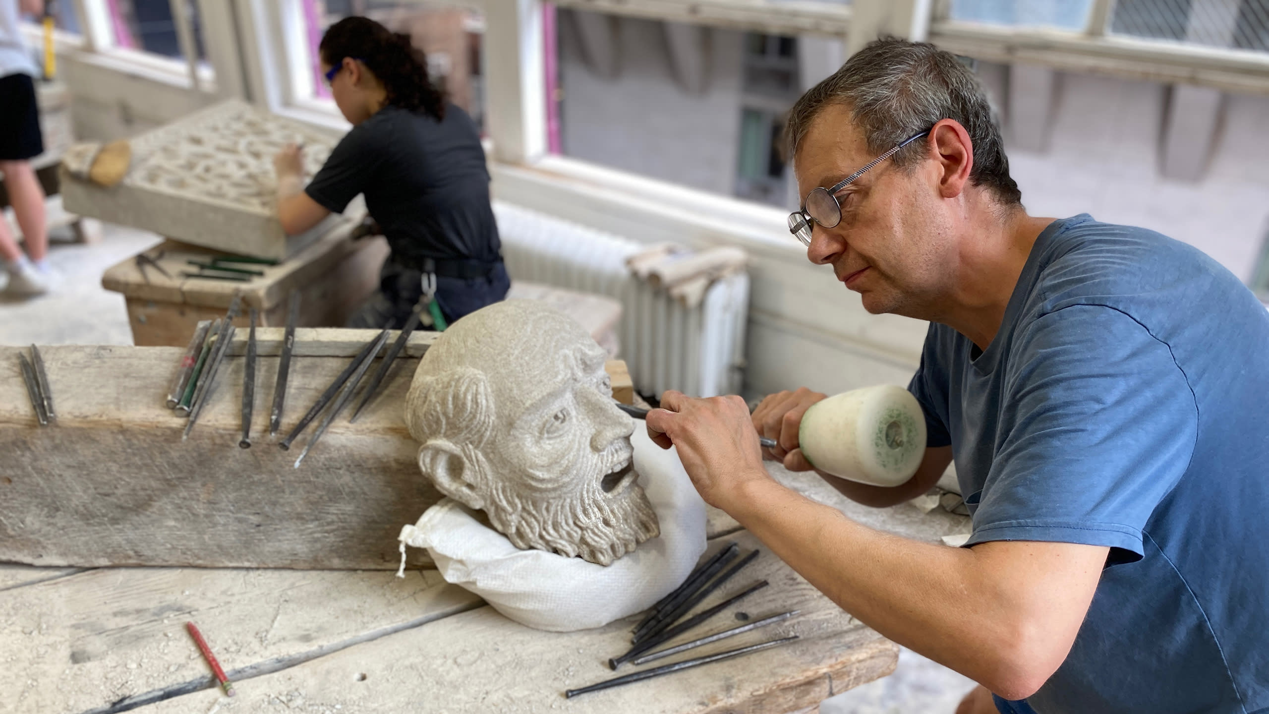 A man carves a detail into a stone statue on a worktable in front of him.