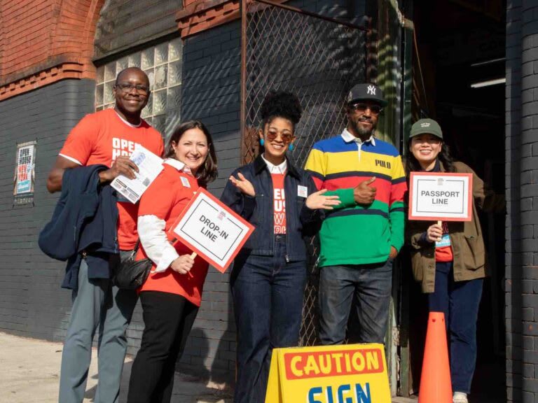 Five smiling people stand outside a brick building. Three wear red shirts and hold signs that help direct visitors.
