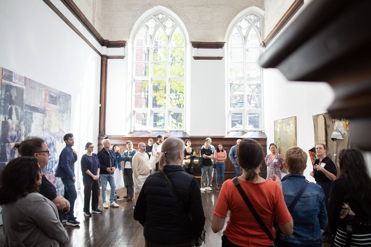 A tour group listens to a docent discuss the paintings on the gallery walls. The space is a high-ceilinged former church with arched gothic windows.