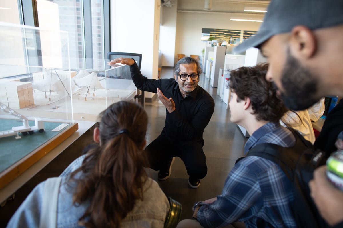 A group of people listen to an engineer discuss the building model on a cabinet next to him.