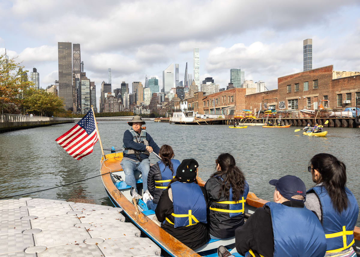 A six-person rowboat departs the dock in Long Island City. The Manhattan skyline is visible in the background.