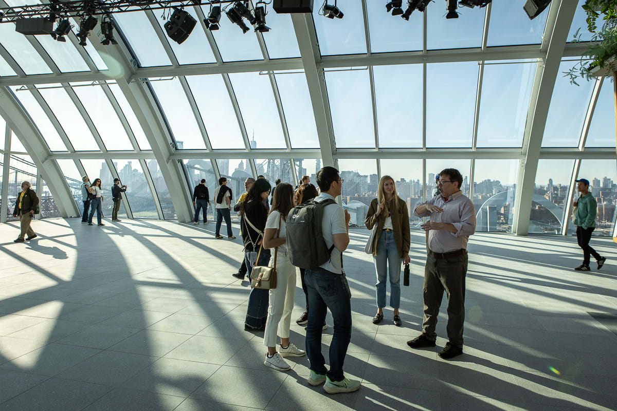 People stand inside a glass atrium overlooking a city skyline and a suspension bridge in the background.