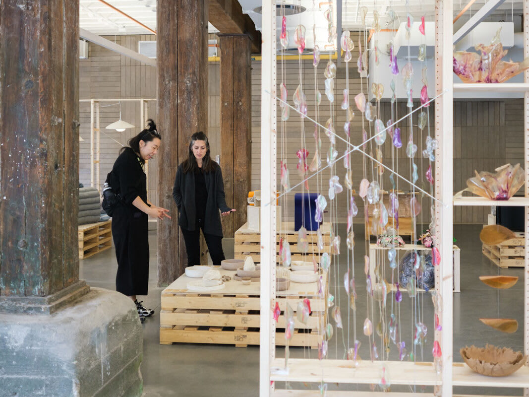 Two people stand in an art gallery with wooden columns, examining ceramic pieces on wooden pallets. Colorful glass or resin ornaments hang on strings in the foreground, partially obscuring shelves with more art objects.