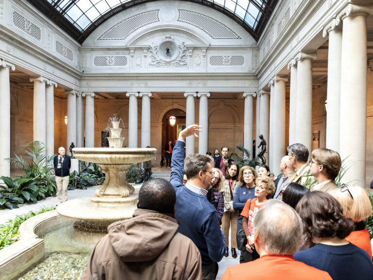 A group of people listens to a guide in a grand indoor courtyard with a fountain, columns, plants, statues, and a glass ceiling. The guide gestures upward, and the group looks attentive.