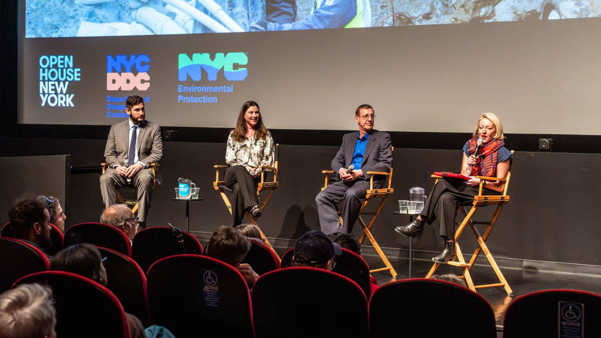 Four people speak on stage at a panel event in a theater, with audience seated and logos on the screen behind them.