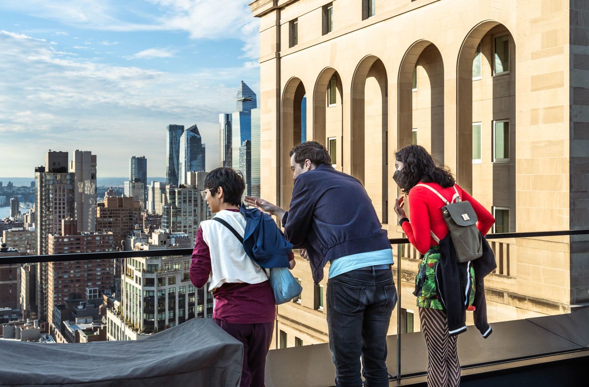 Three people look over a glass balcony on the roof of a building in Midtown. The Manhattan skyline facing west is visible around and in front of them.