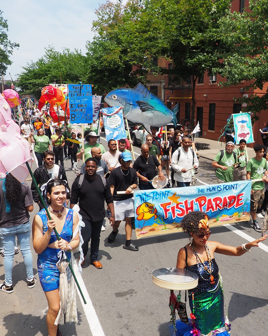 Participants march in the 2023 Fish Parade, carrying colorful signs, drums, and a large puppet shaped like a blue fish.. 