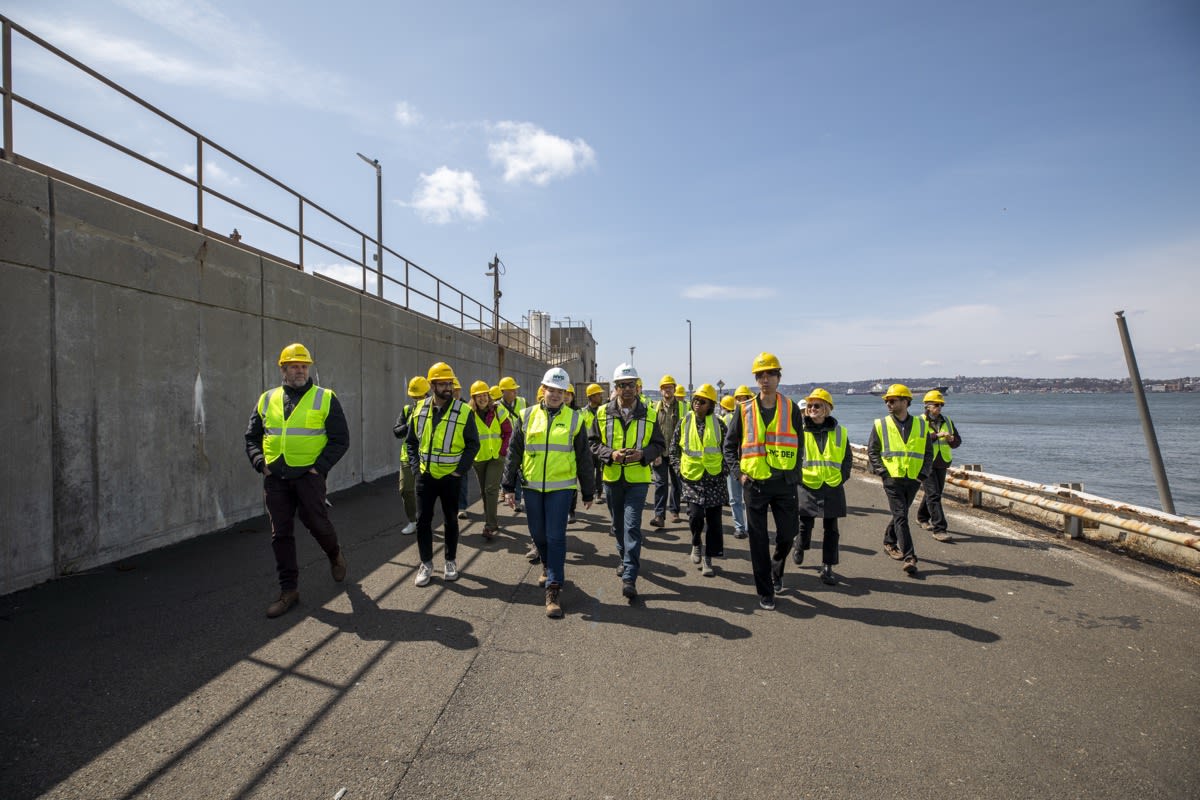 A group of workers in safety vests and hard hats walk by the water near a concrete wall on a sunny day.