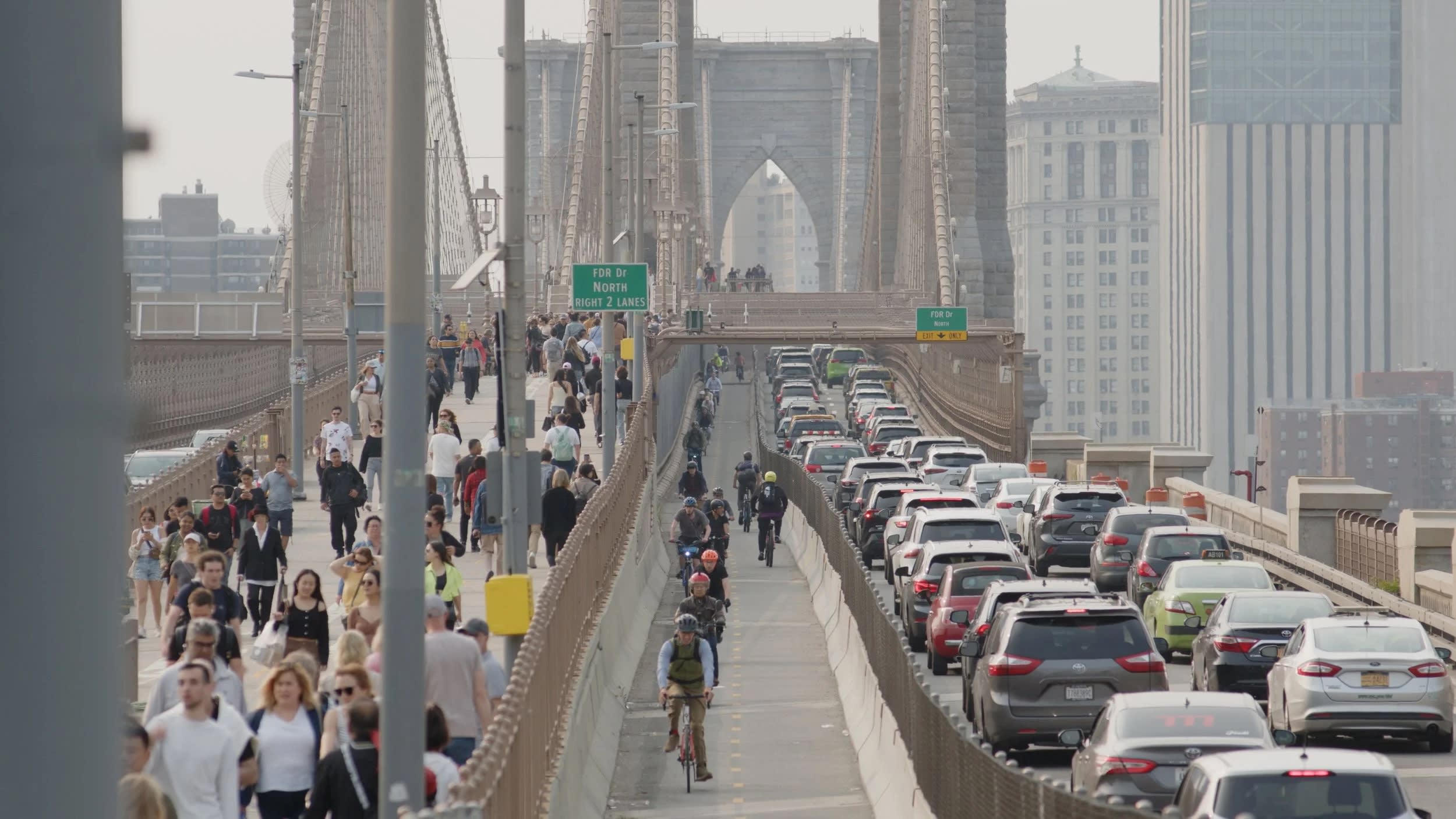 Crowds of people walk and bike alongside heavy car traffic on a bridge in an urban setting.