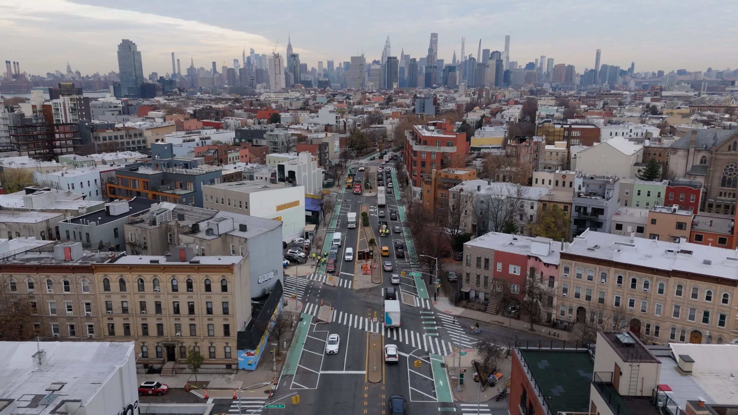 A busy city street with cars, surrounded by buildings, and a distant skyline under a cloudy sky.
