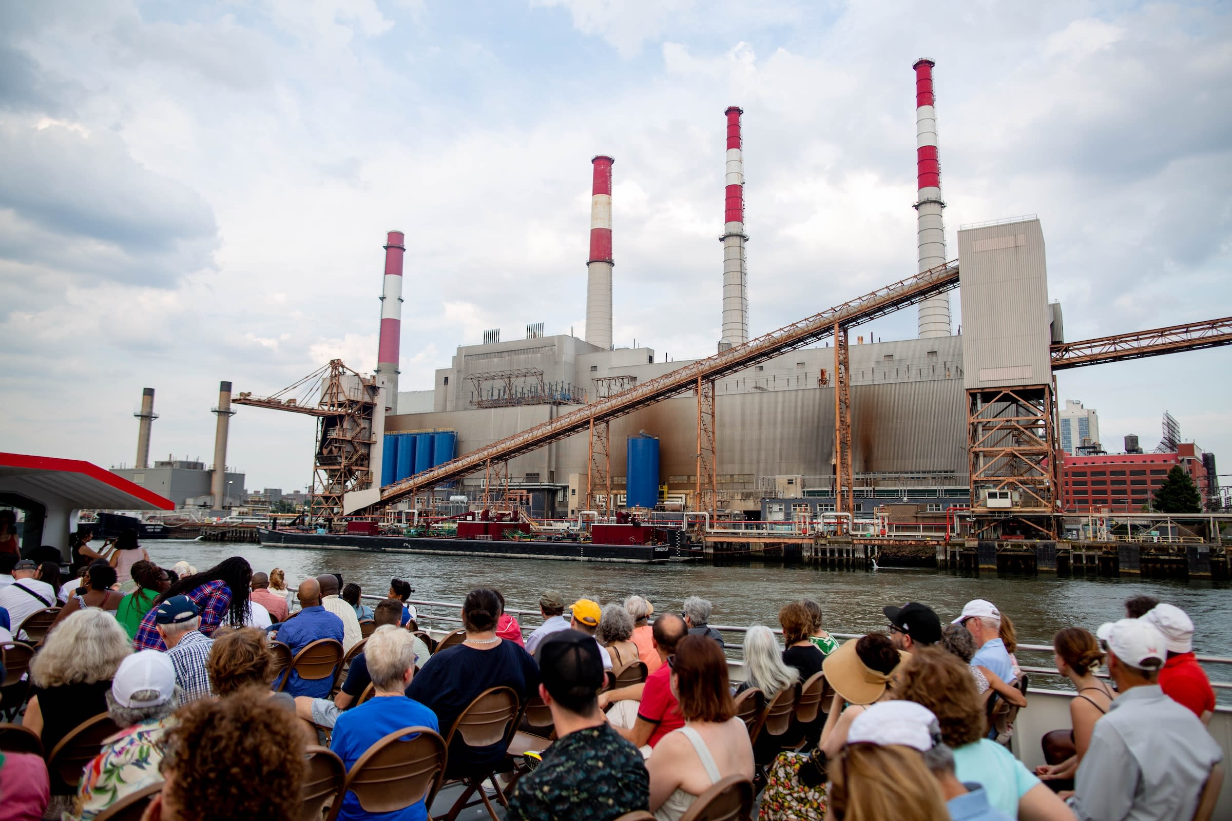 A large audience sits by a river, facing an industrial power plant with tall chimneys and red-striped stacks.