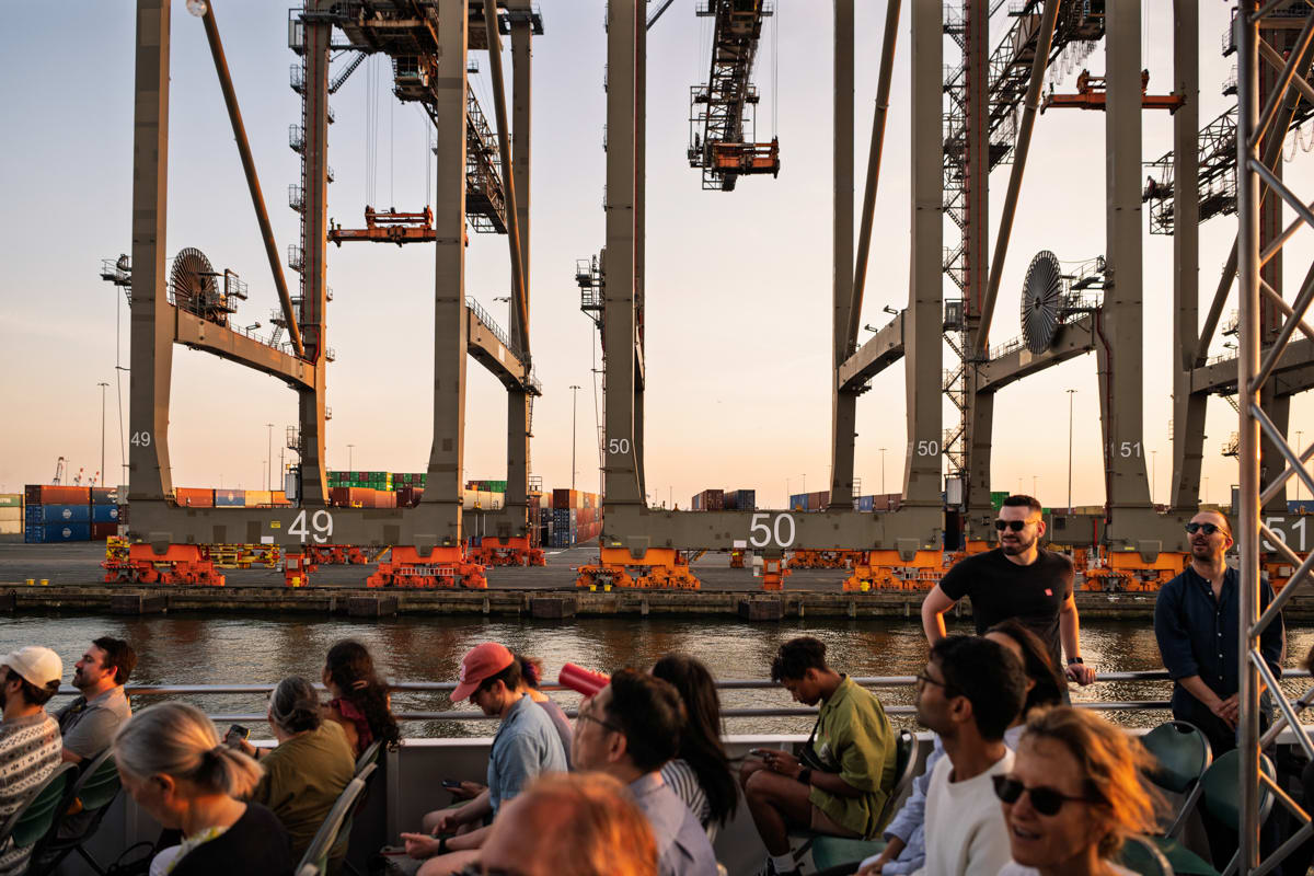 People sit on a boat by large shipping cranes at a port, with containers and water in the background at sunset.
