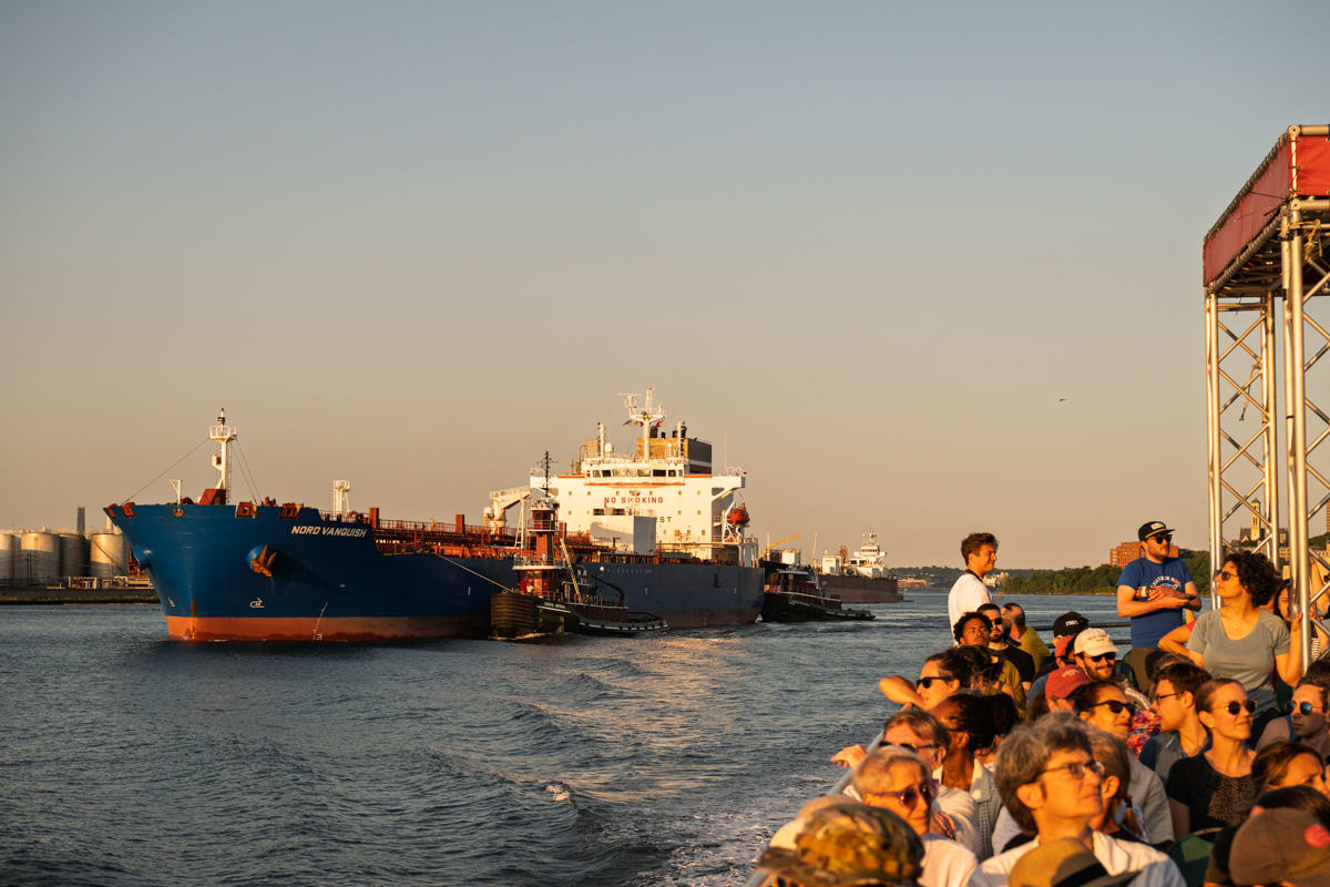 A large blue cargo ship sails near a crowd of people on a boat during sunset, with calm water and clear sky.