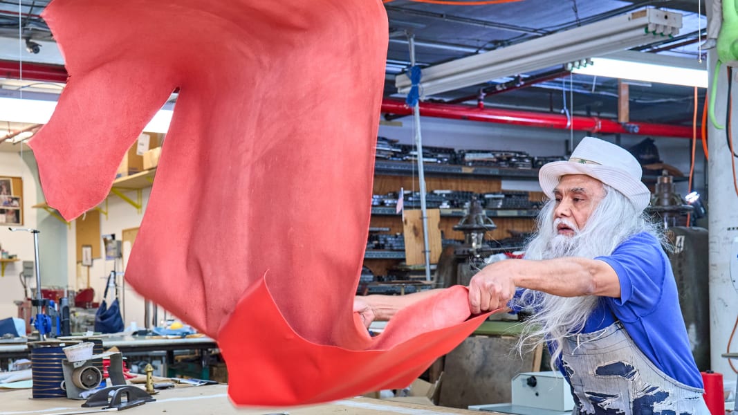 An elderly man with a long white beard tosses a large red leather hide in a workshop.
