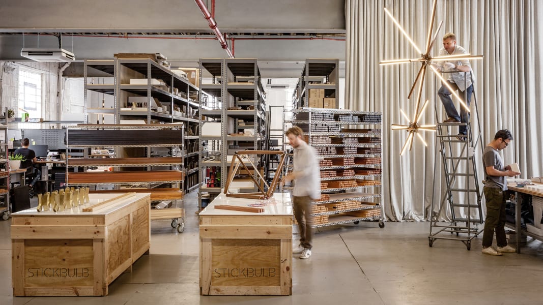 Four people work in a modern workshop with shelves, wood, and bright, geometric light fixtures.