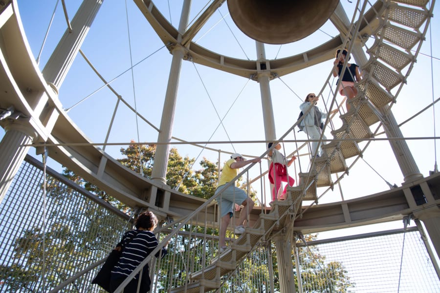 People climbing a spiral staircase inside a tall, open metal structure with a large bell above them.