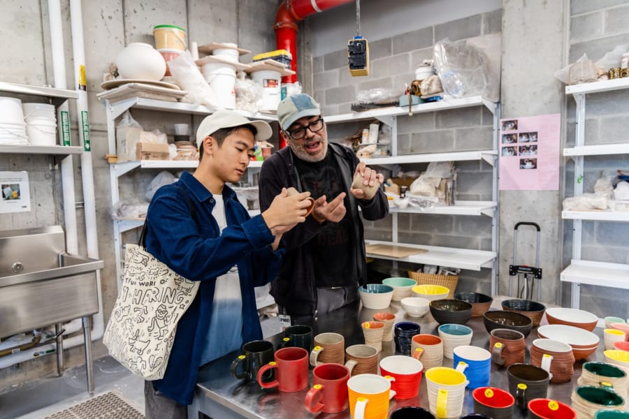 Two people examine pottery cups on a table in a studio filled with shelves of ceramics and art supplies.