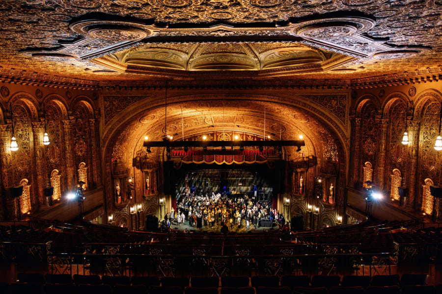 Ornate theater interior with elaborate ceiling, balcony seating, and a large party gathered on stage.