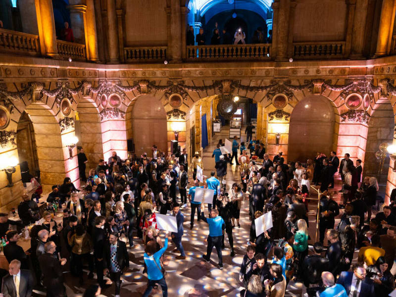 A large crowd gathers in an ornately lit hall with dancers in blue shirts performing in the center.