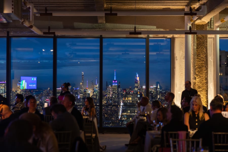 People dining indoors at night with large windows overlooking a city skyline illuminated by lights.