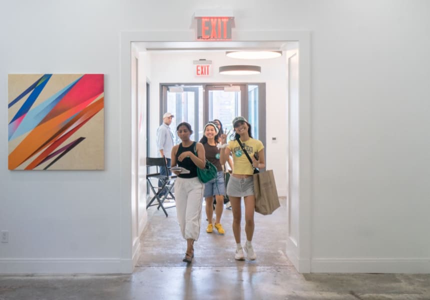 Three young women smiling and waving as they walk through a hallway, with a man standing in the background.