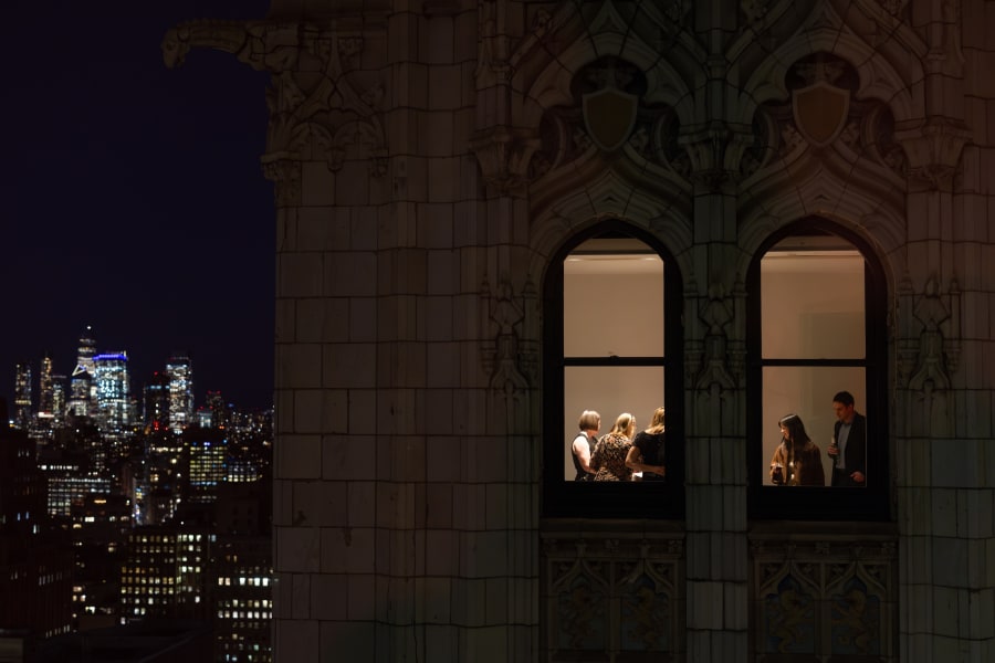 A group of people stand and talk inside a lit room at night, viewed through two arched windows of a tall building, with a city skyline visible in the background.