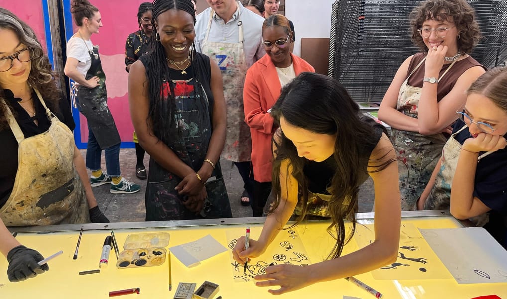A group of people watch a woman draw on a lit yellow table covered with art supplies and sketches..